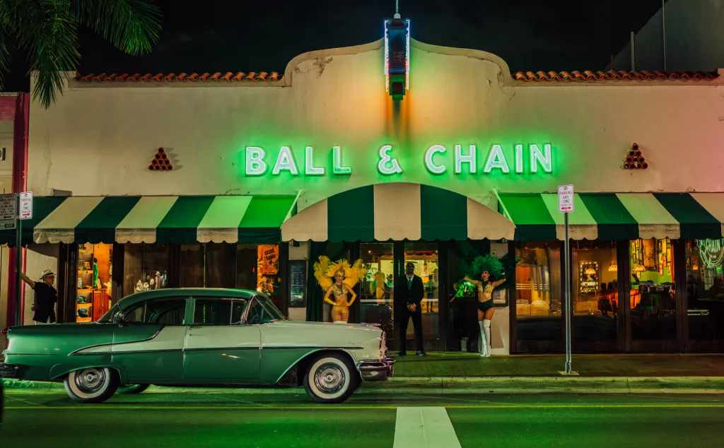 The exterior of Ball & Chain bar at night, featuring green neon signs, a classic car, and two dancers in feathered costumes.