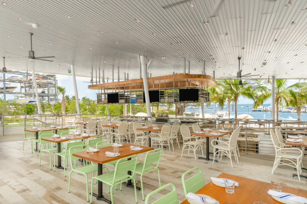 An outdoor restaurant patio at Bayshore Club overlooking a marina with boats and palm trees.