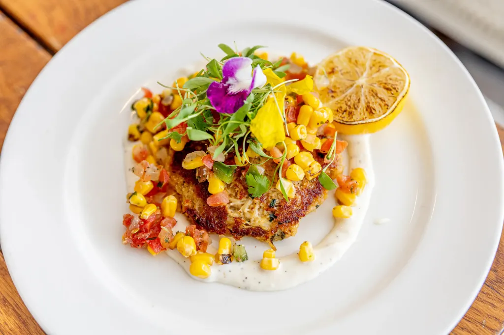 Crab cake with corn salsa, a purple edible flower, and a charred lemon slice on a white plate.