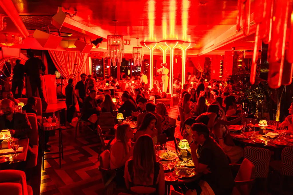 A lively restaurant interior bathed in red light, with patrons seated at tables and a performer on a stage in the background.