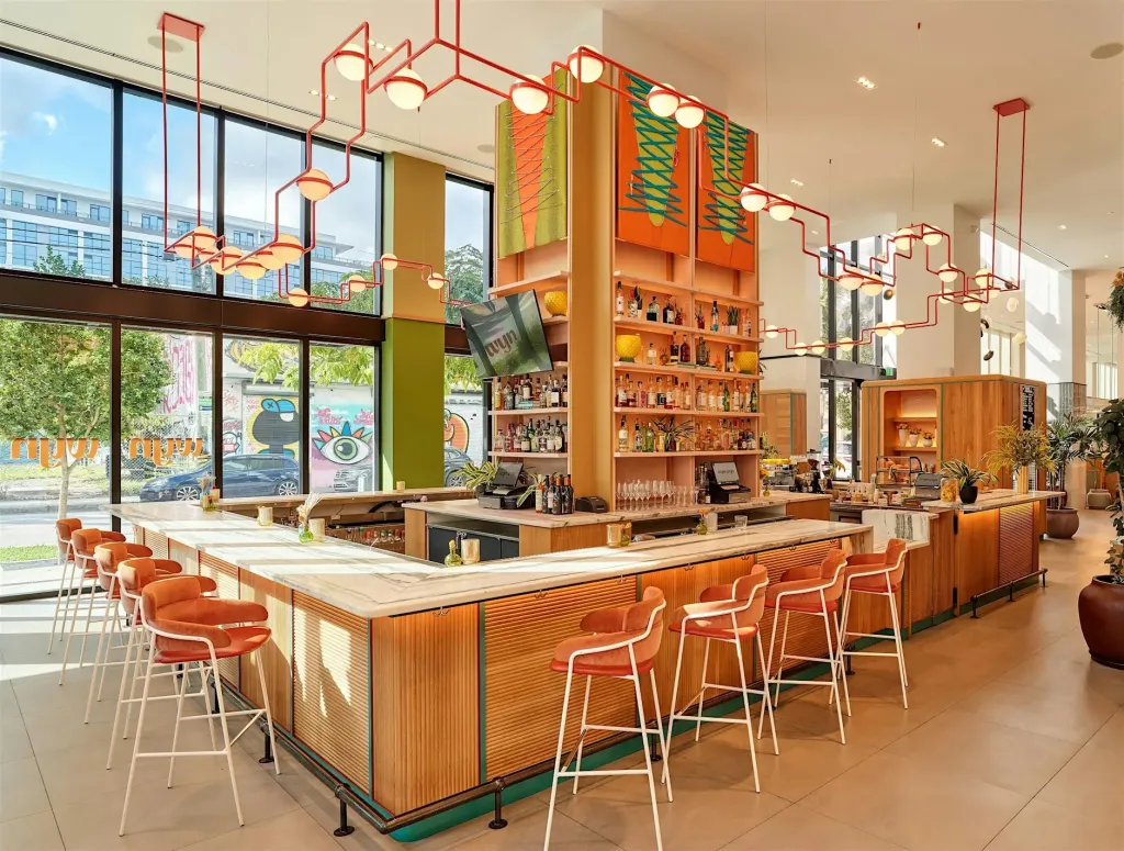 A contemporary bar and lounge with a wooden bar, coral-colored chairs, and large windows looking out onto a street with colorful graffiti.