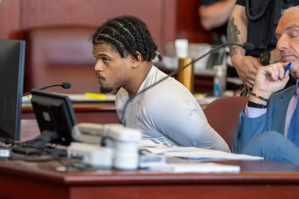 Halley Tejada, with braided hair and a goatee, is seen in a courtroom wearing a light-colored long-sleeved shirt, handcuffed, looking to his left, with a man in a blue suit next to him.