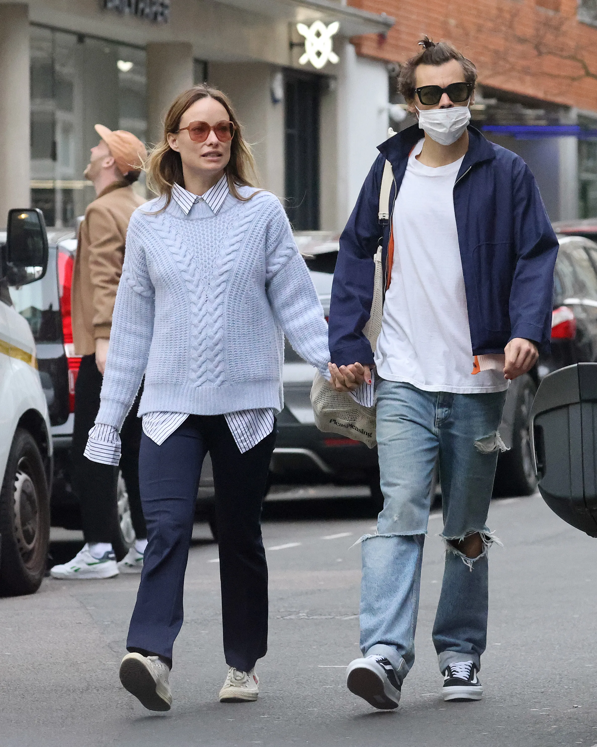 Harry Styles, wearing a face mask and sunglasses, holding hands with Olivia Wilde, wearing a light blue sweater and sunglasses.