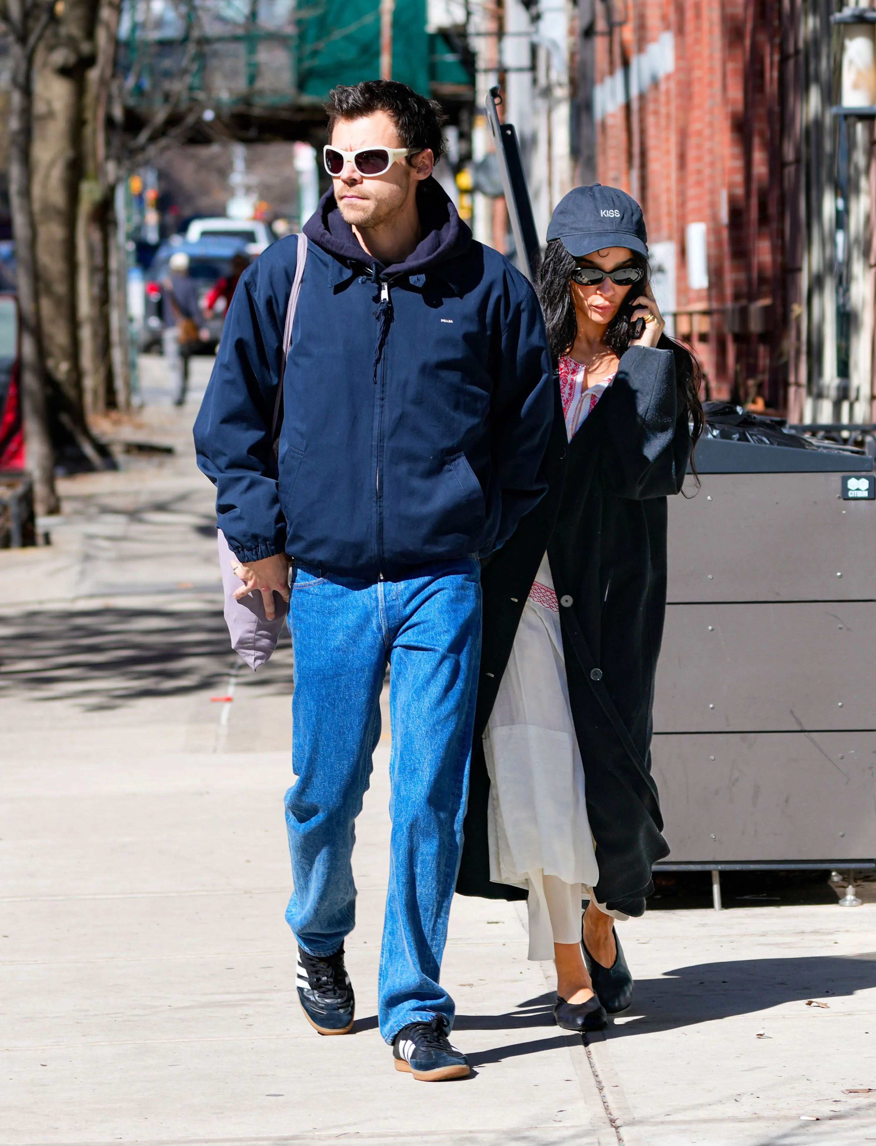 Harry Styles wearing a blue jacket and jeans with Zoe Kravitz in a black hat and coat, walking on a city street.