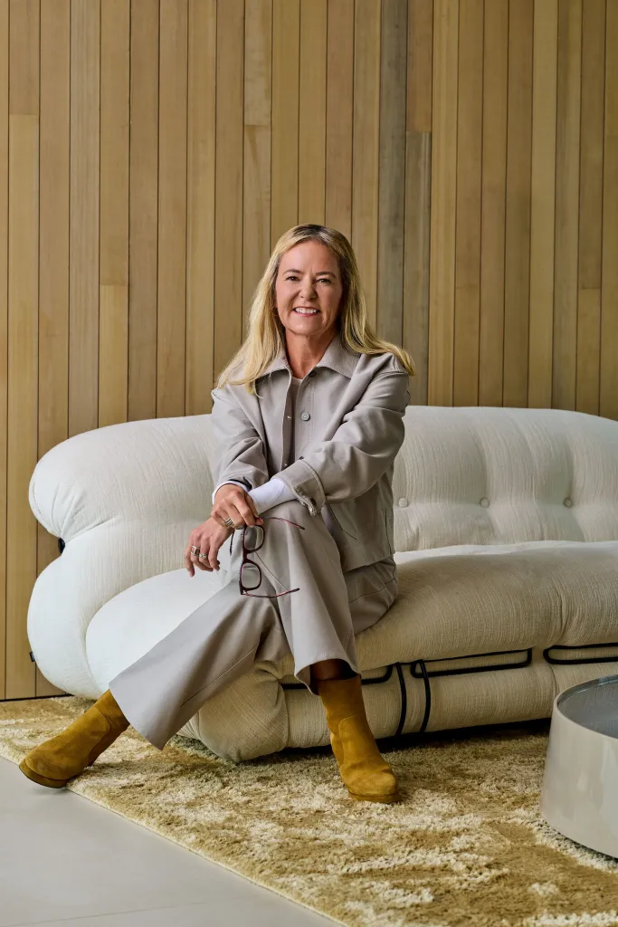 Heidi O'Neill, with blonde hair, sitting on a white sofa and holding eyeglasses.