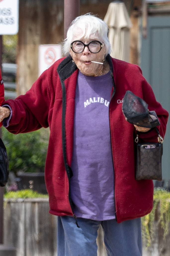 Shirley MacLaine in a red jacket, purple "Malibu" t-shirt, sweatpants, black glasses, white hair, and holding a lollipop in her mouth.