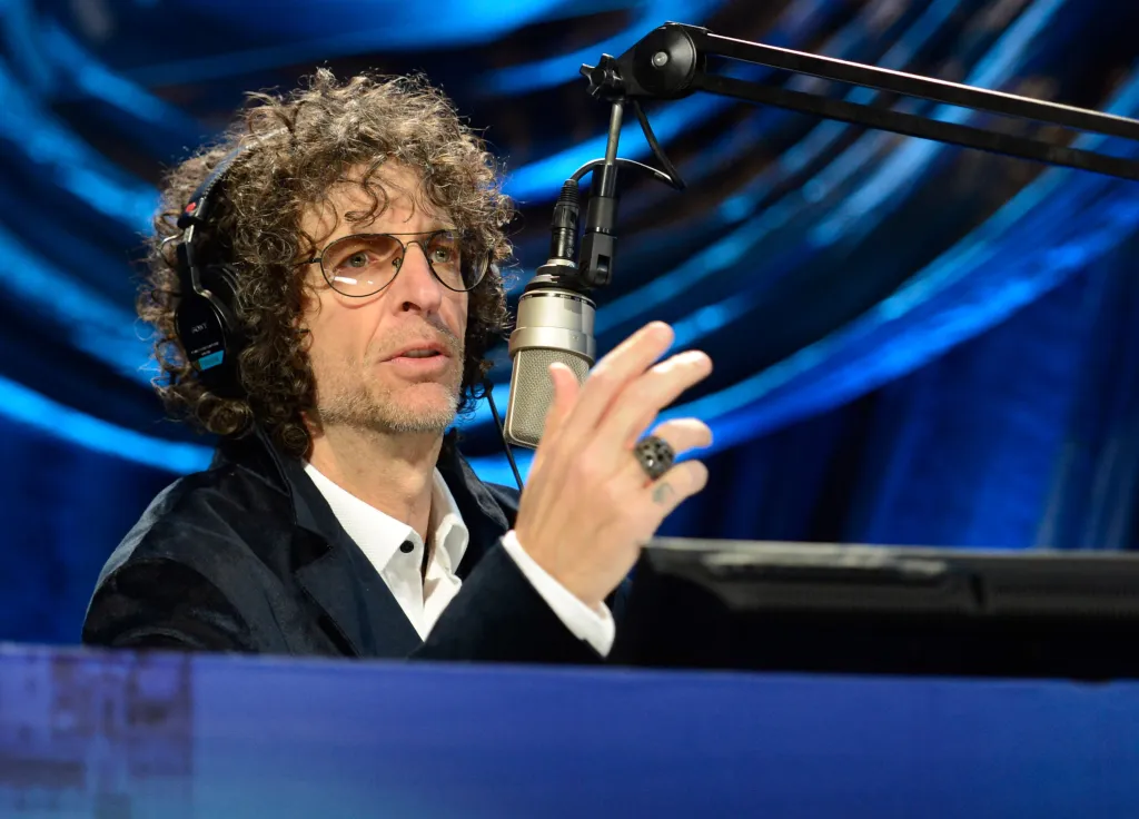Howard Stern speaking onstage at his birthday bash, wearing headphones and gesturing with his hand.