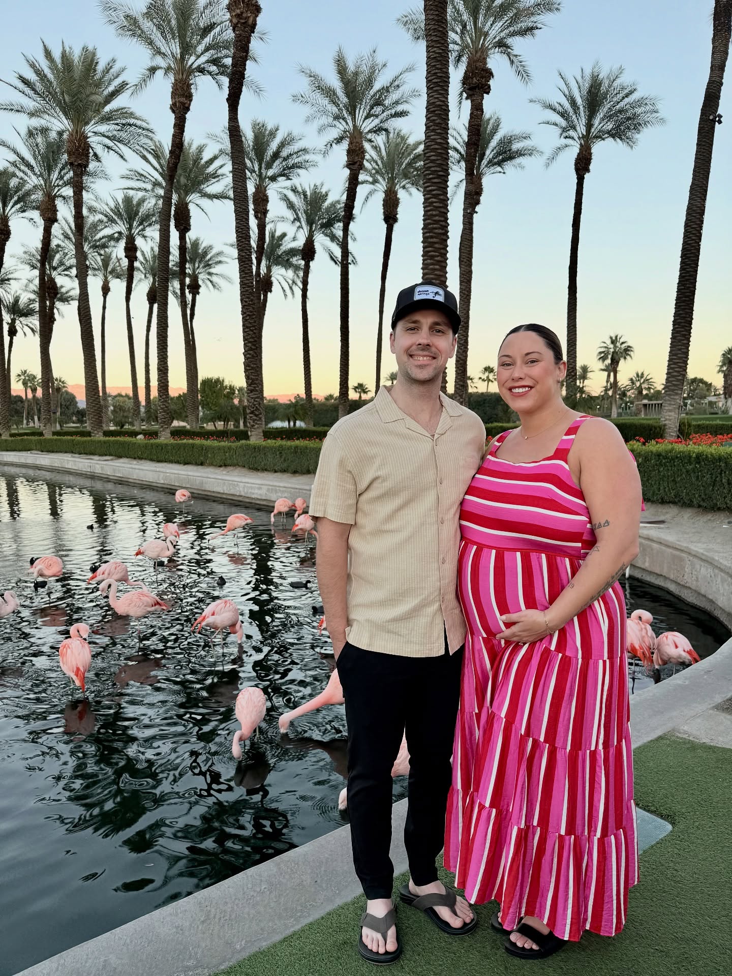 A pregnant woman and her husband smiling next to a pond filled with flamingos.