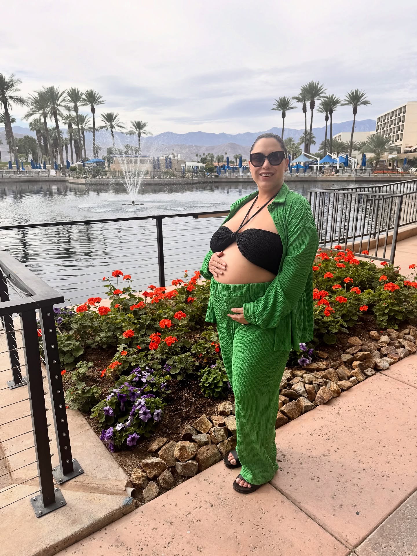 Alaina Scott in a green top and matching pants, showing off her baby bump by a lake with a fountain.