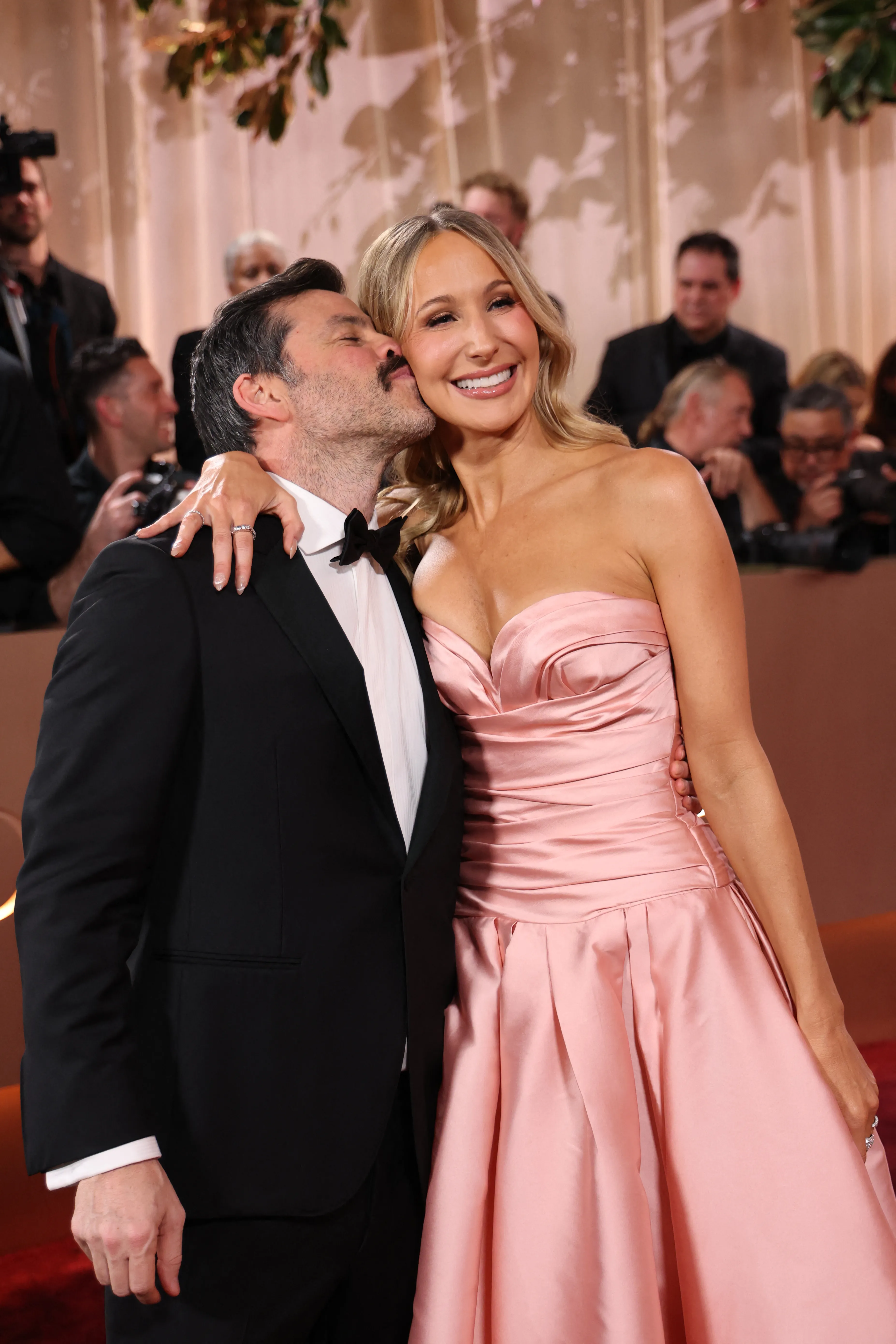 Nikki Glaser in a pink strapless gown with Chris Convy, who is kissing her cheek, at the Golden Globe Awards.