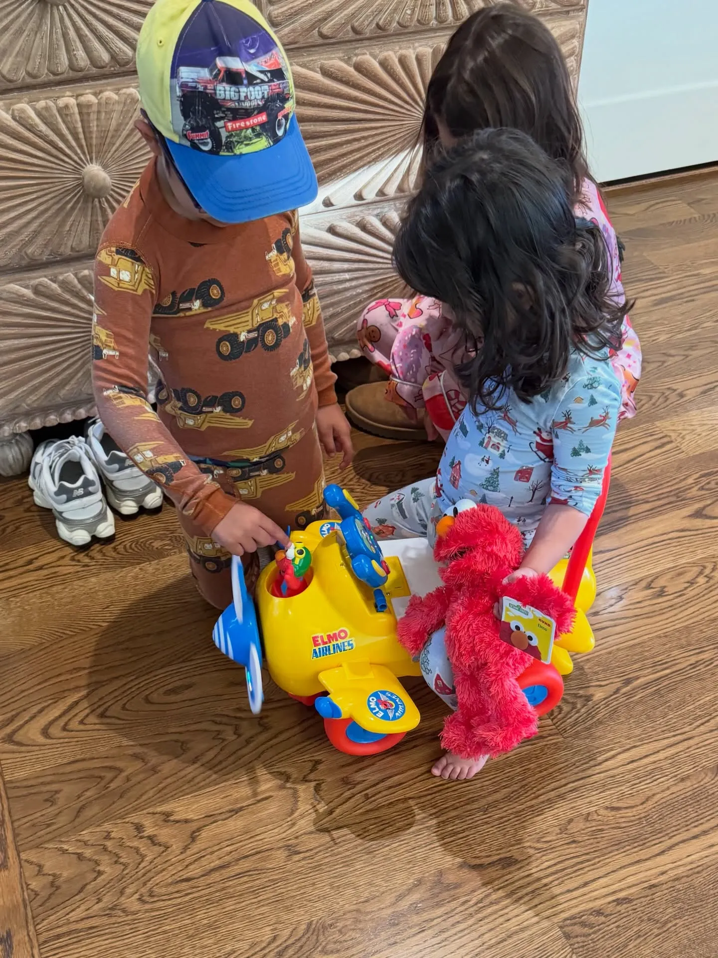 Three children playing on a wooden floor with a yellow Elmo Airlines toy plane and a red Elmo doll.