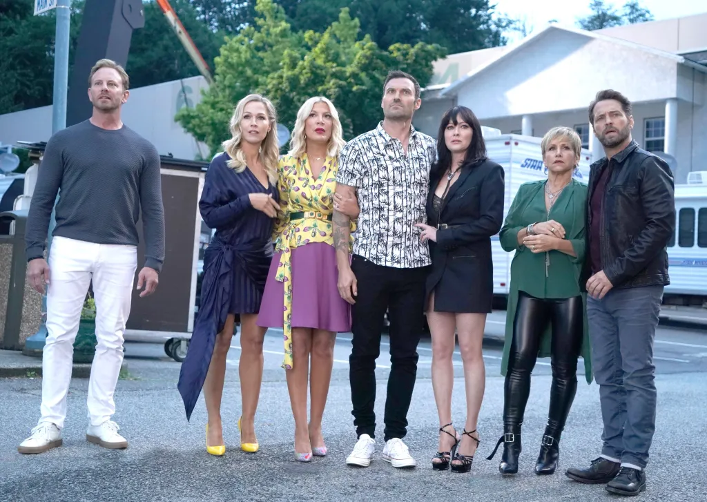 Cast shot of Ian Ziering, in a grey sweatshirt, white pants, Jennie Garth, in a black dress, Tori Spelling, in a yellow printed top and purple skirt, Brian Austin Green, In a black and white patterned shirt and black pants, Shannen Doherty, in a black jacket Gabrielle Carteris, in a green top and black leather pants, and Jason Priestley, in a black leather jacket and jeans, in "BH90210."