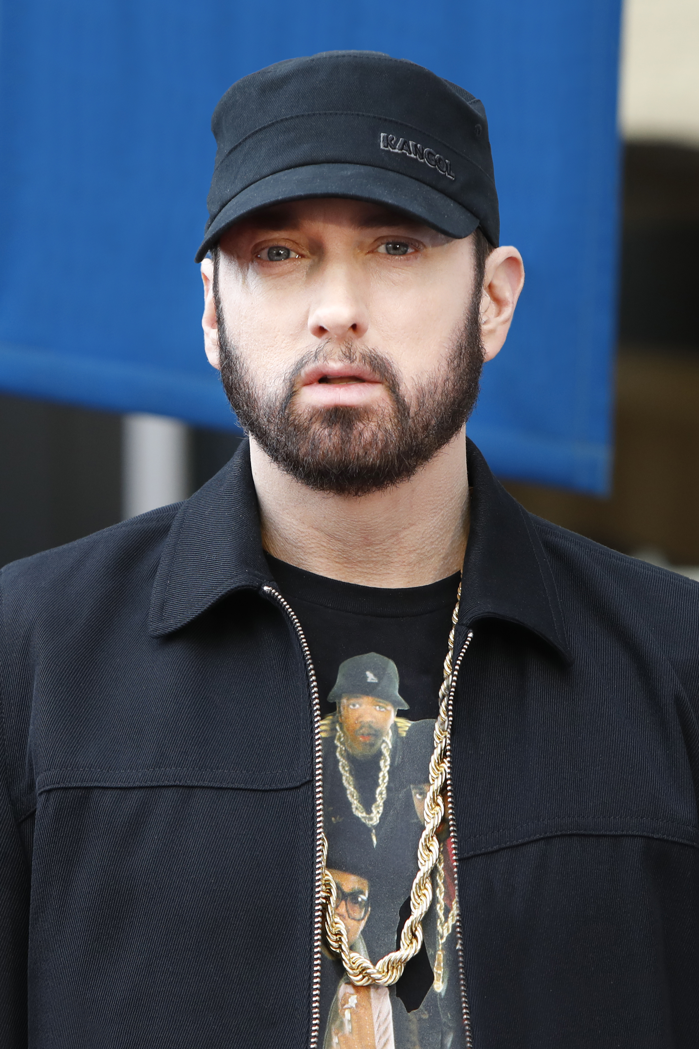 Eminem, wearing a black Kangol hat, black jacket, and a gold chain over a graphic t-shirt, stands for a photo.