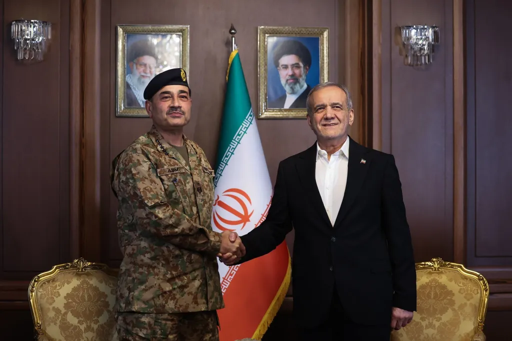 Masoud Pezeshkian shaking hands with Marshal Asim Munir, with the Iranian flag and portraits of Ayatollah Khomeini and Ayatollah Khamenei in the background.