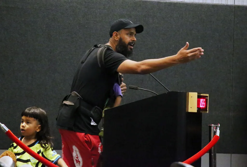 A man speaks at a podium with a timer displaying "0:55", while a young child stands in the foreground.