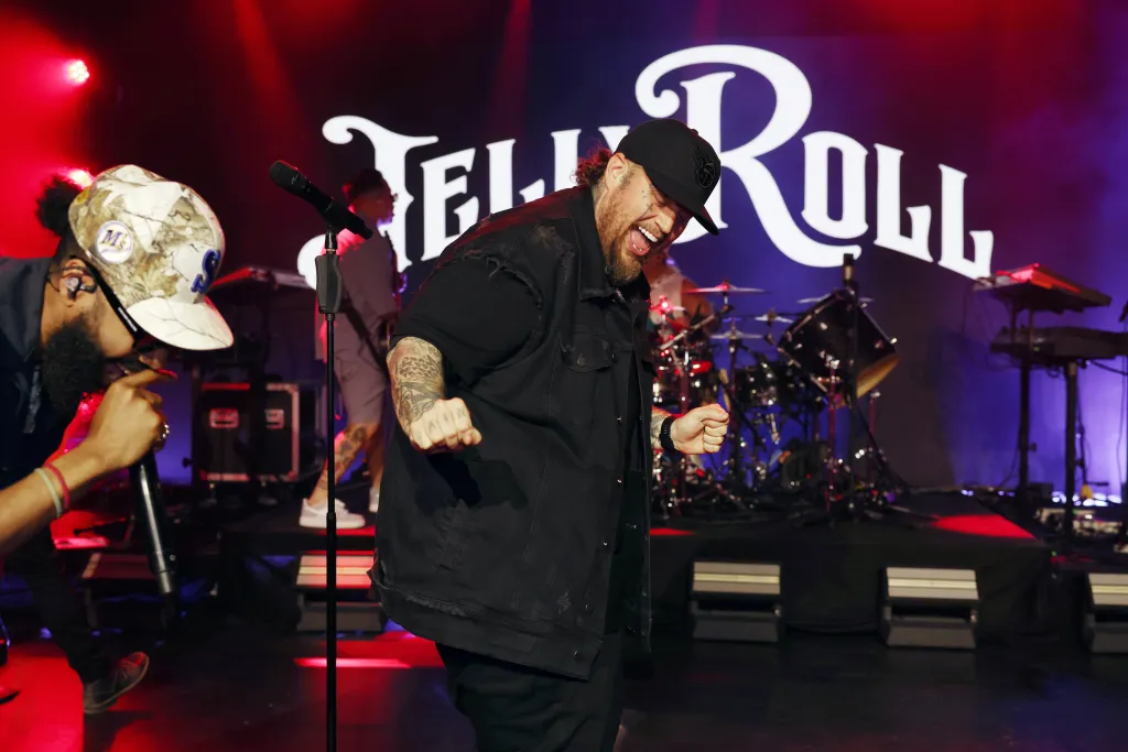 Jelly Roll performing on stage, wearing a black vest and hat, smiling with his mouth open, with a large "Jelly Roll" logo in the background.