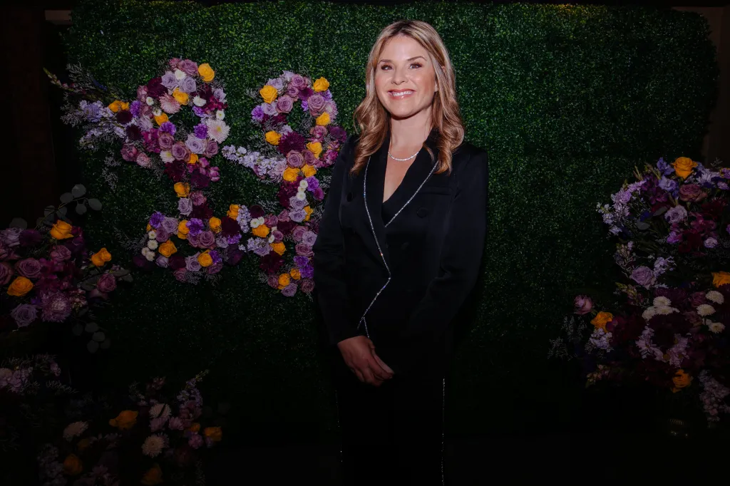 Jenna Bush Hager smiles in a black suit with sparkling lapels and a diamond necklace, standing before a green wall adorned with purple, yellow, and white flowers arranged to form "90".