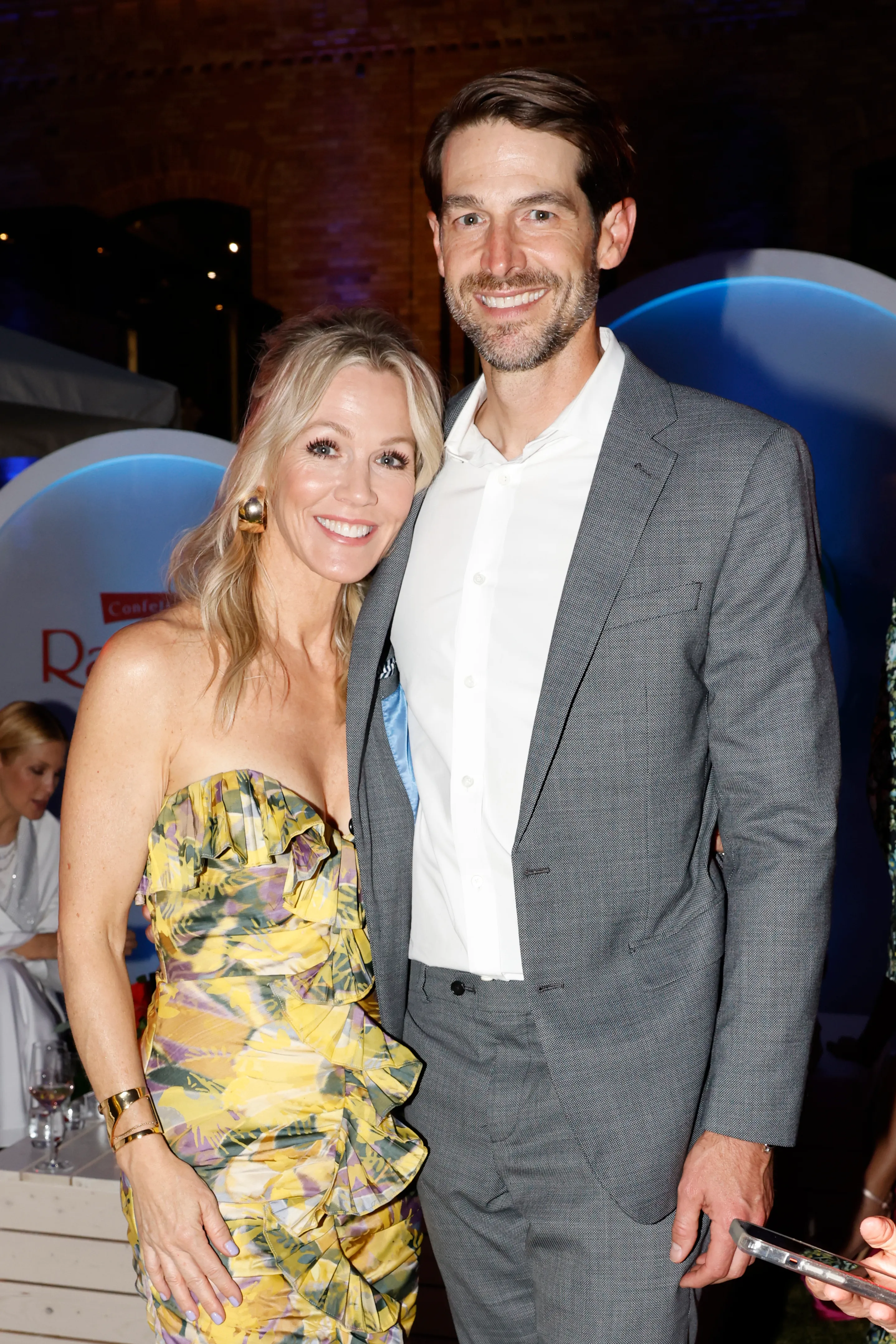 A man in a gray suit and a woman in a yellow and purple patterned dress smile, leaning into each other.