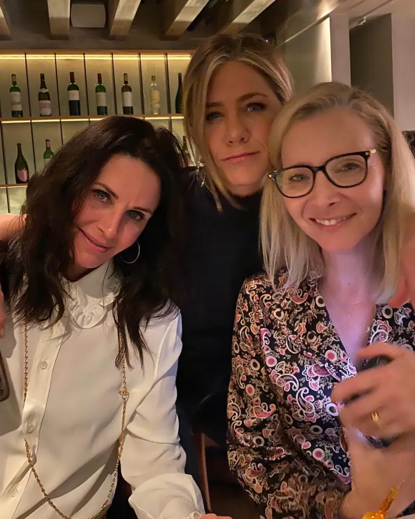 Jennifer Aniston, Courteney Cox, and Lisa Kudrow posing together in a bar with wine bottles on shelves behind them.
