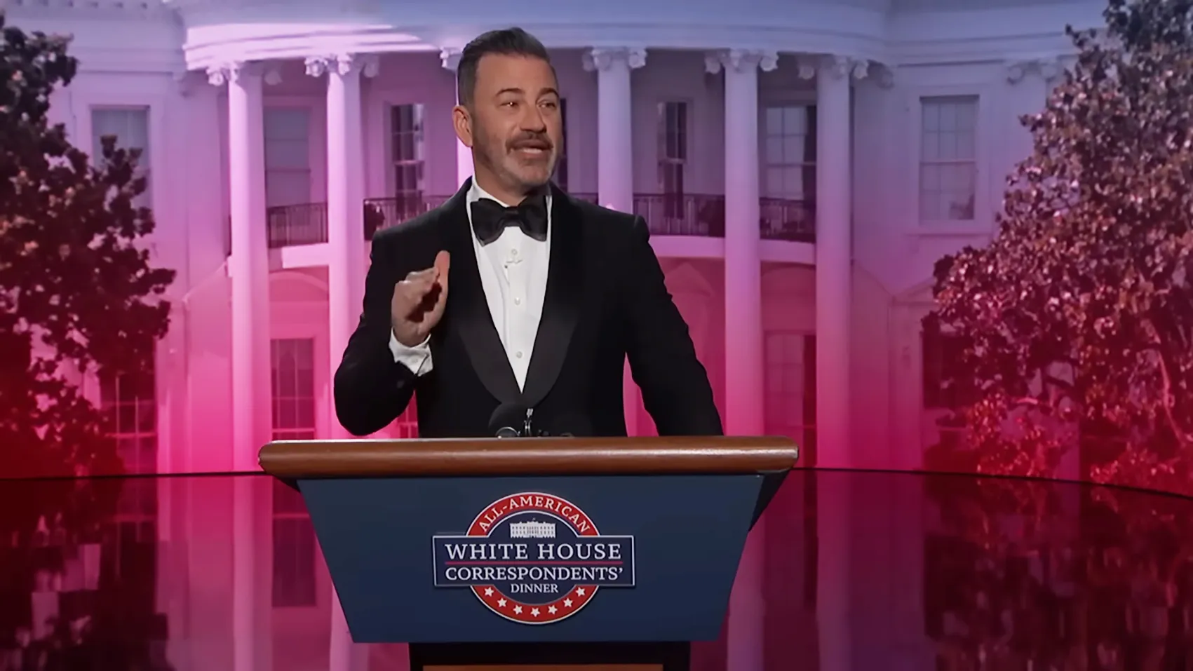 Jimmy Kimmel at the White House Correspondents' Dinner podium with the White House in the background.