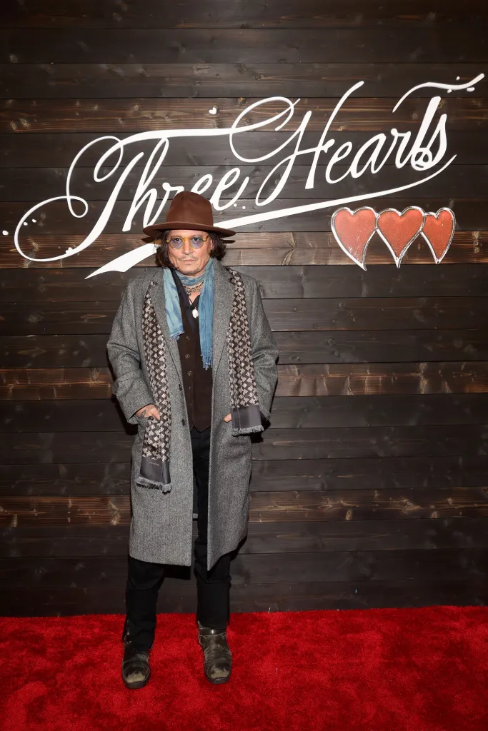 Johnny Depp standing on a red carpet in front of a dark wooden wall with "Three Hearts" written in white script and three red hearts next to it.