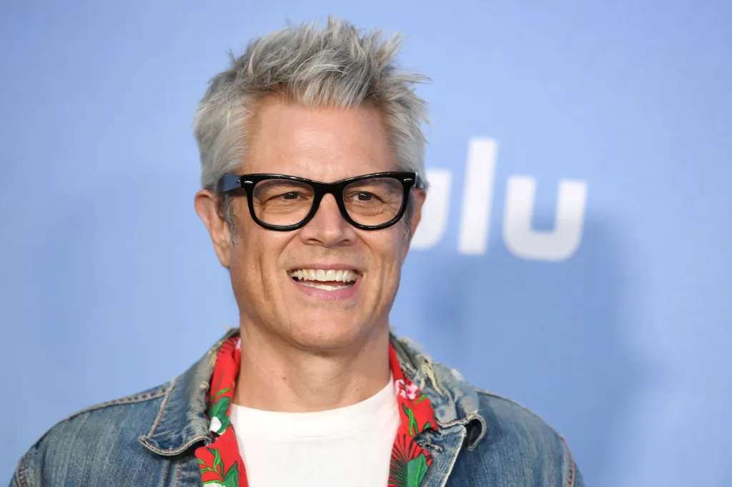 Johnny Knoxville in black glasses, a white t-shirt, and a denim jacket with a red shirt underneath, smiling against a blue background with the white "Hulu" logo.