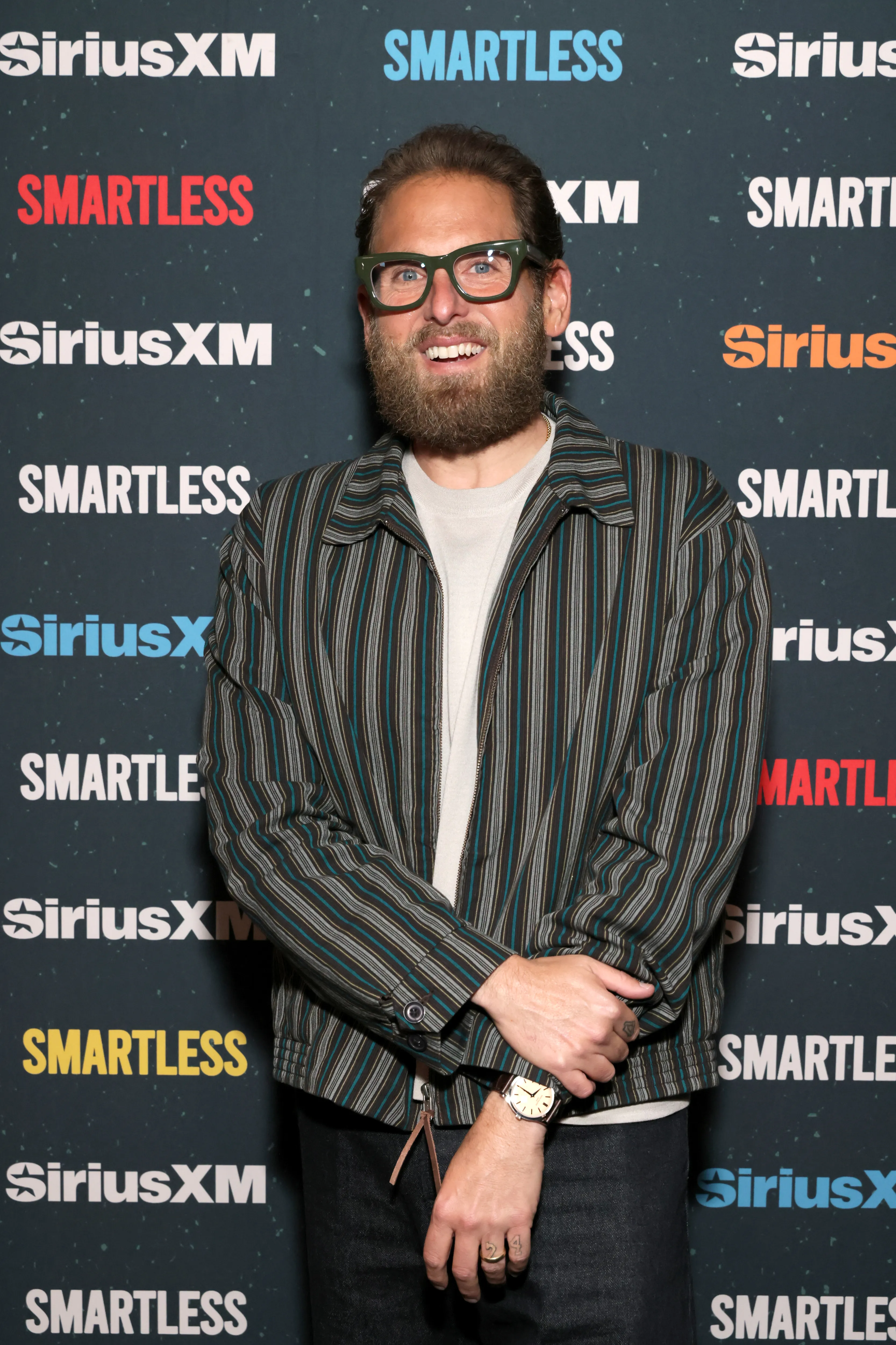 Jonah Hill in a striped jacket and green glasses, smiling at a "SiriusXM SmartLess" event.