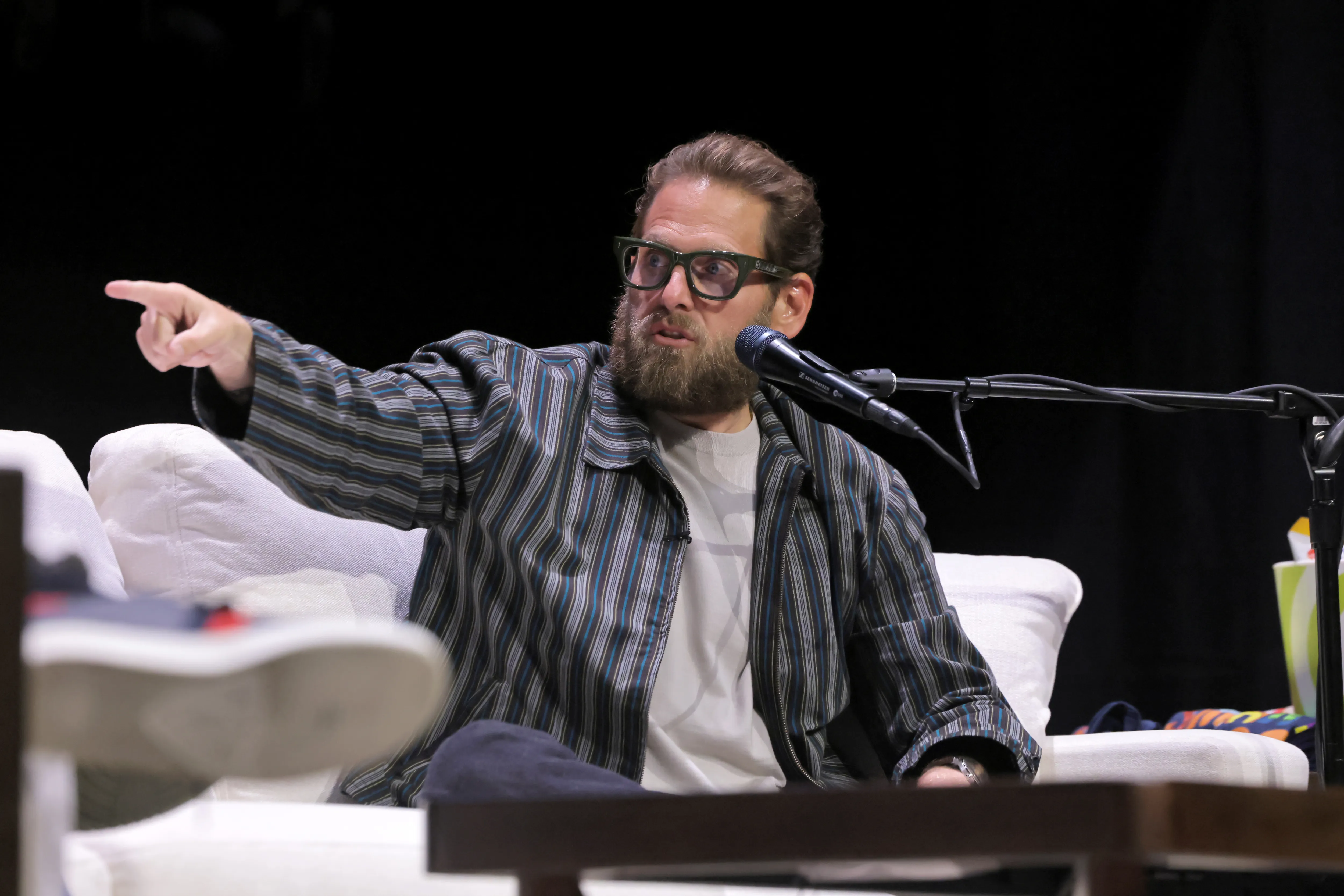 Jonah Hill speaking onstage, wearing glasses and pointing his right hand.