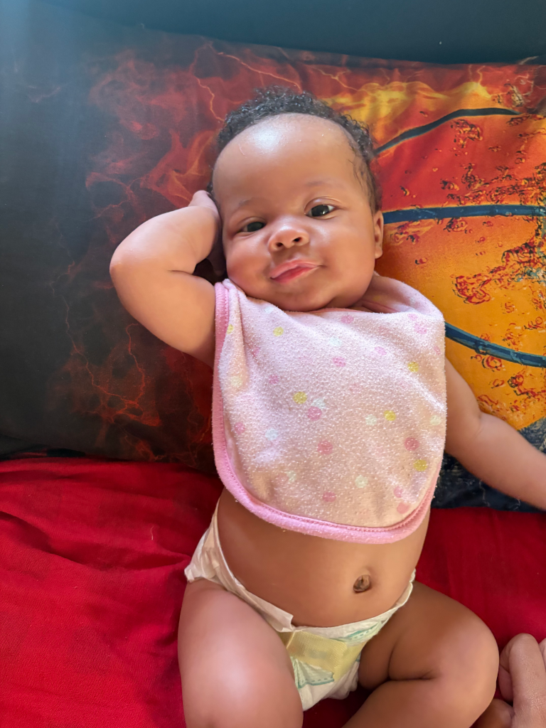 Kaori Patterson Moore, a baby girl, lying on a red bedspread with a patterned pillow, wearing a pink bib and a diaper.