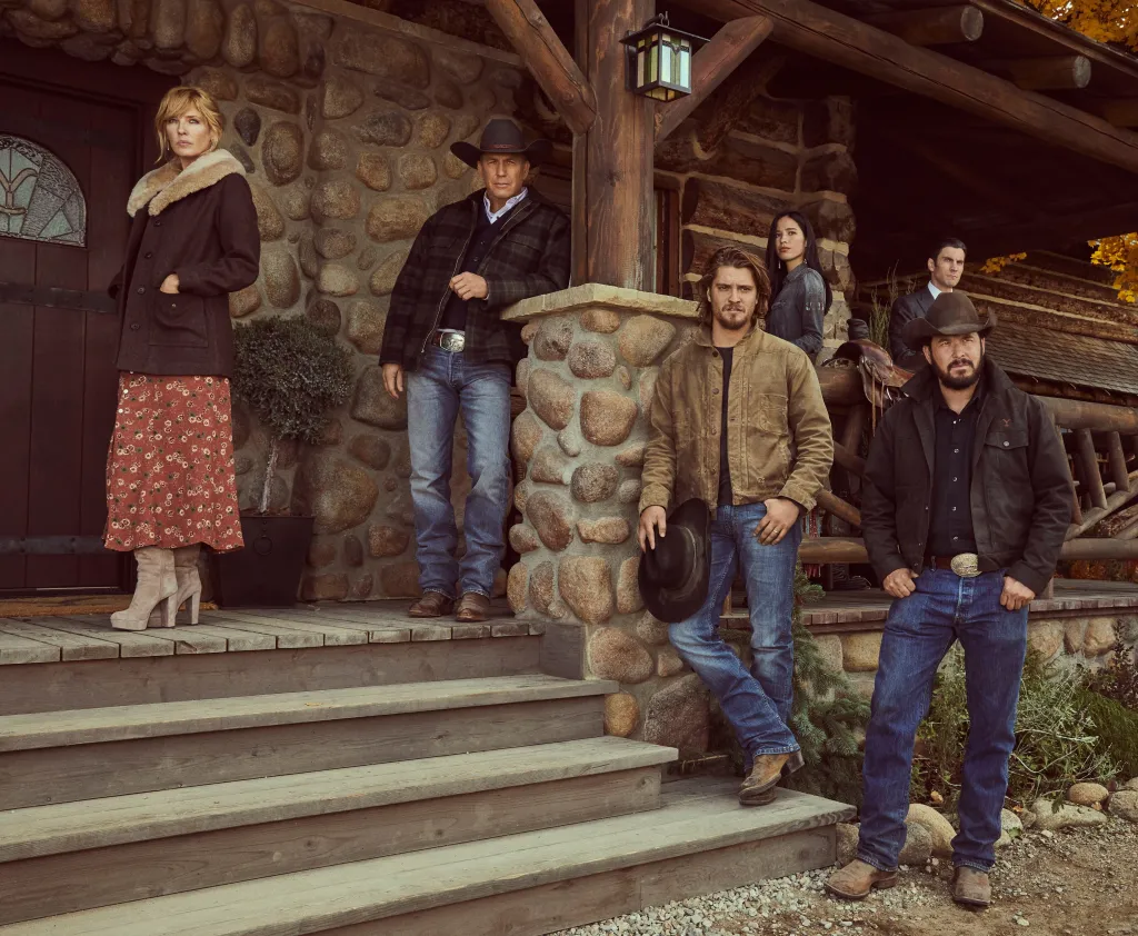 Promotional still from the television show Yellowstone, featuring six actors on the porch of a rustic stone and wood building.