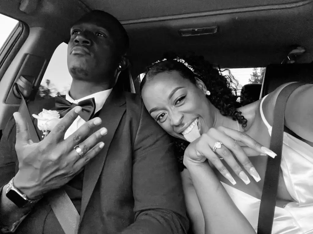 Black and white photo of a man in a tuxedo and a woman in a white dress, both in a car.