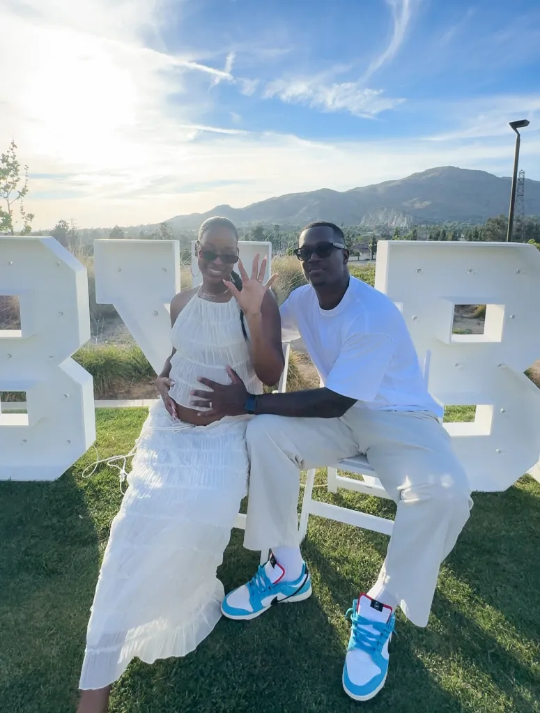 A pregnant woman and a man in matching white outfits sit together outdoors, in front of large white letters that spell "BKB," with a mountain range in the background.