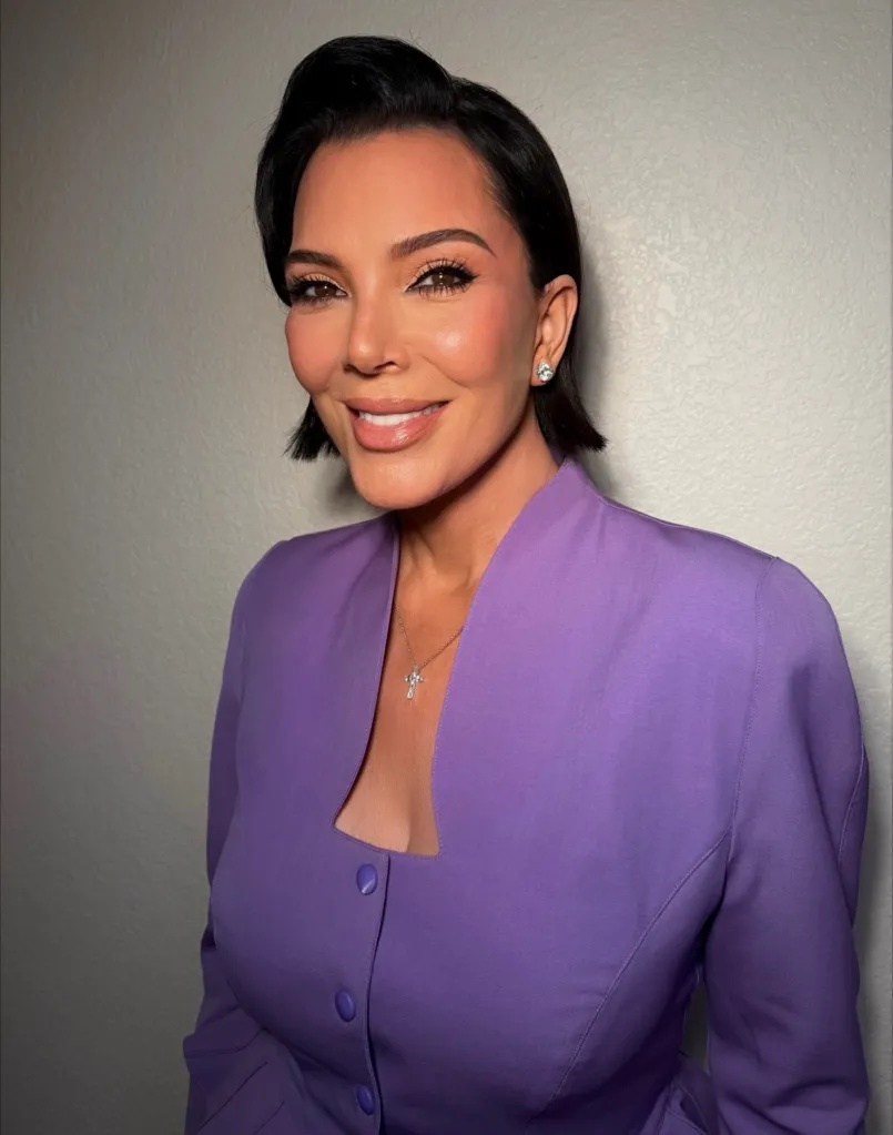 Kris Jenner smiles at the camera, wearing a purple dress and a cross necklace.