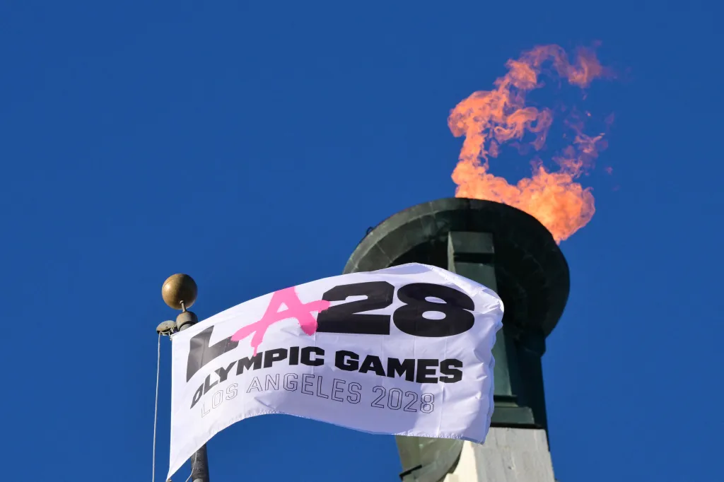 The LA28 Olympic cauldron lit with a flame, with a banner for the Los Angeles 2028 Olympic Games in the foreground.