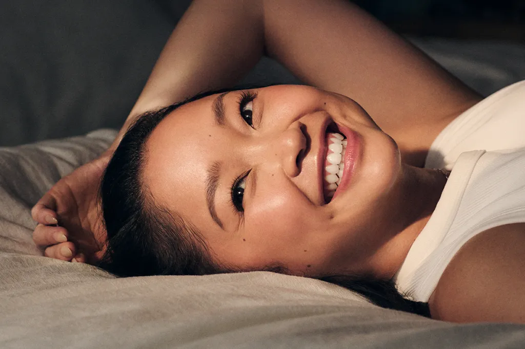 Lana Condor smiling and lying on her back, looking directly up at the camera.