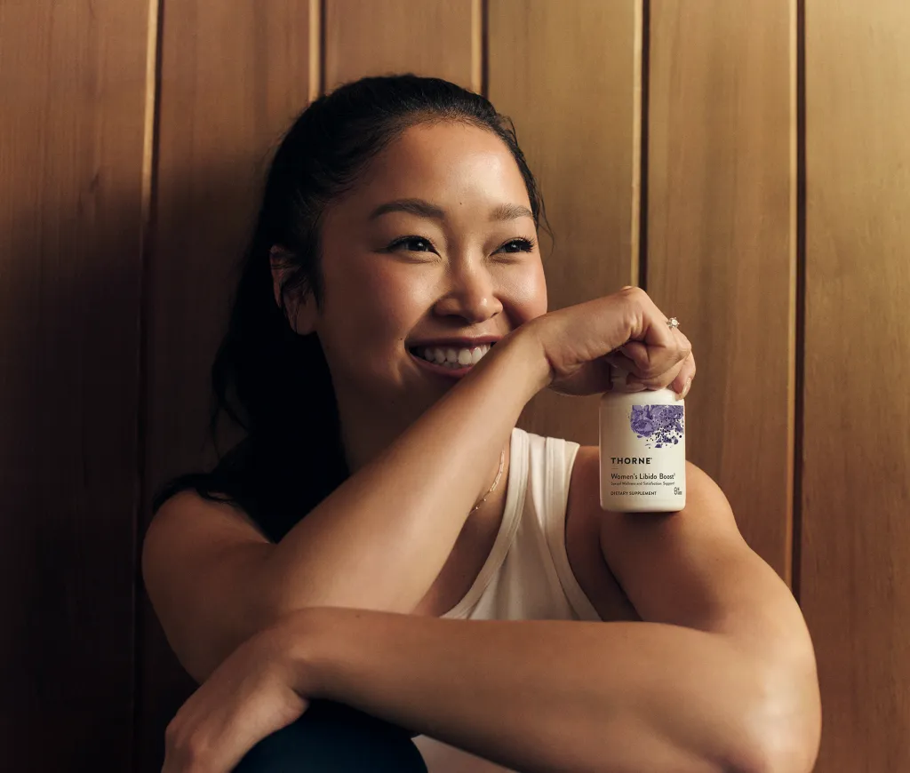 Lana Condor smiling and holding a bottle of Thorne Women's Libido Boost.