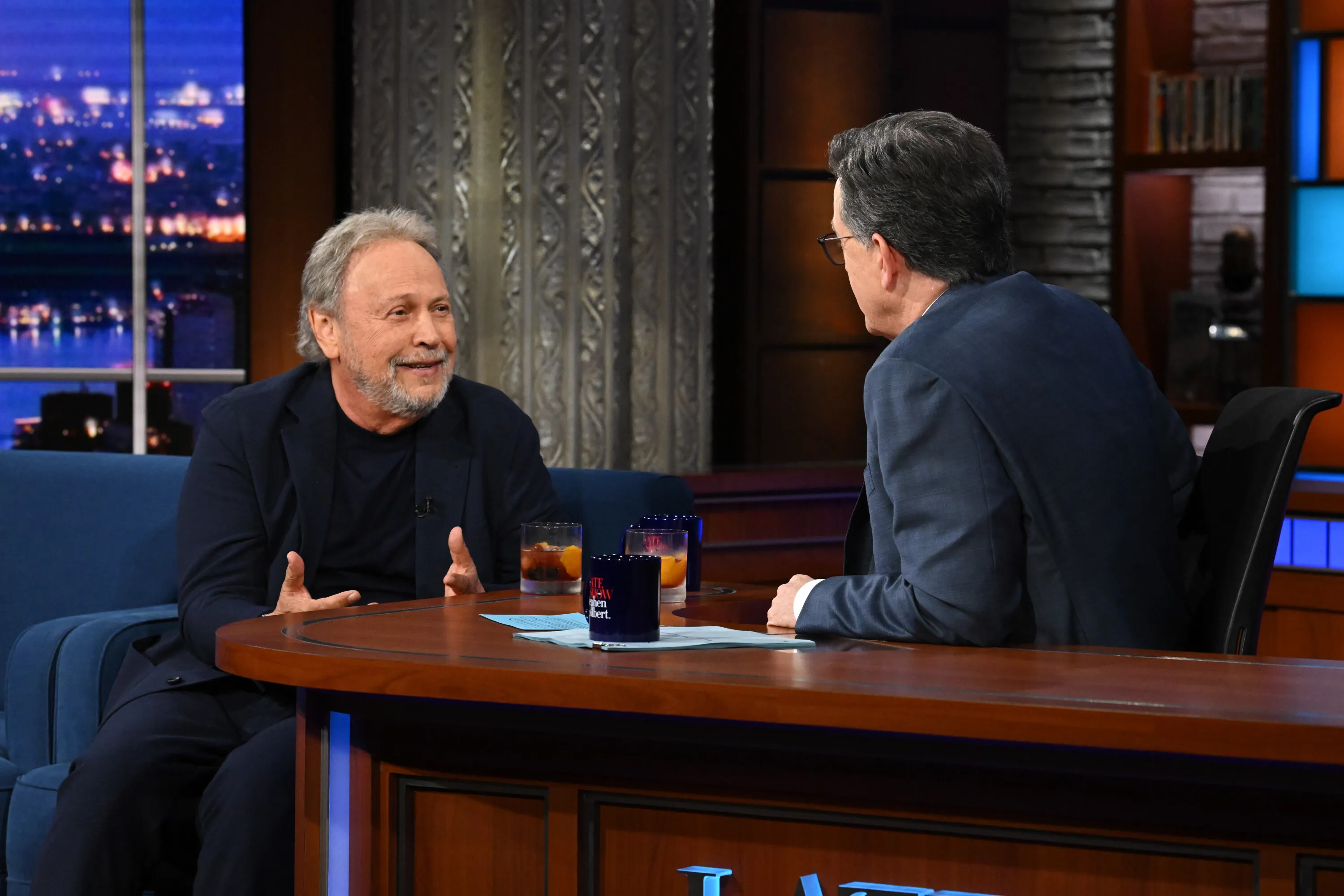 Billy Crystal and Stephen Colbert sit across from each other at a desk on The Late Show.