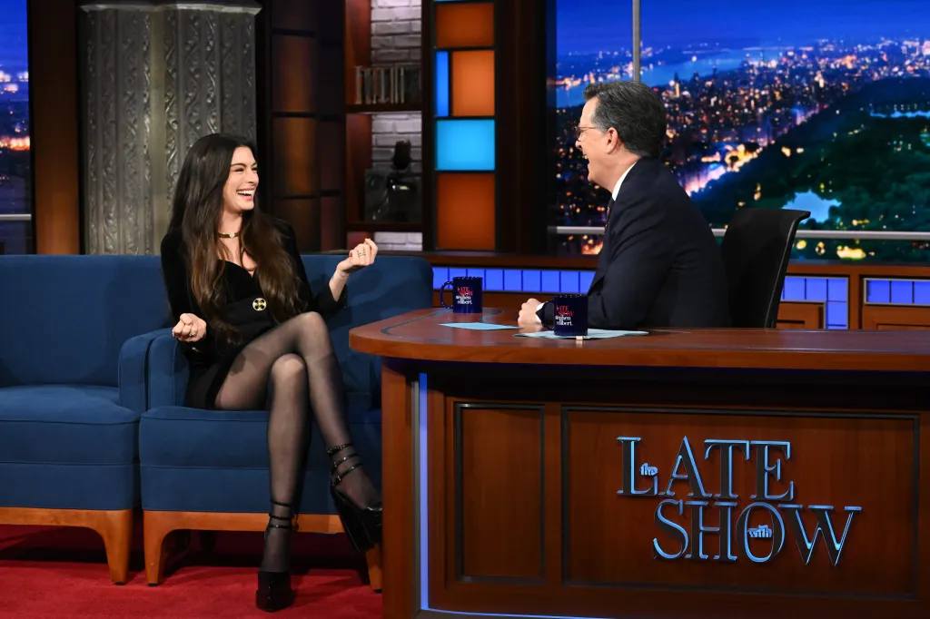 Anne Hathaway sits on a blue couch, laughing with Stephen Colbert, who sits behind a desk.