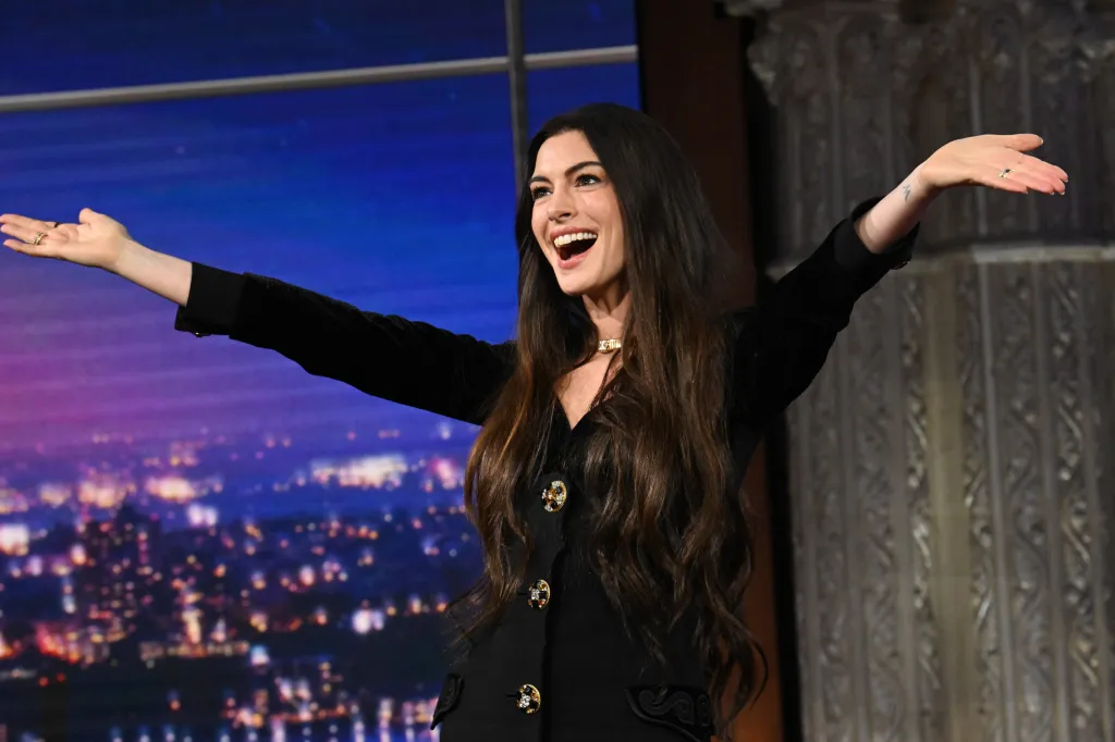 Anne Hathaway smiling with her arms outstretched during an appearance on The Late Show with Stephen Colbert.