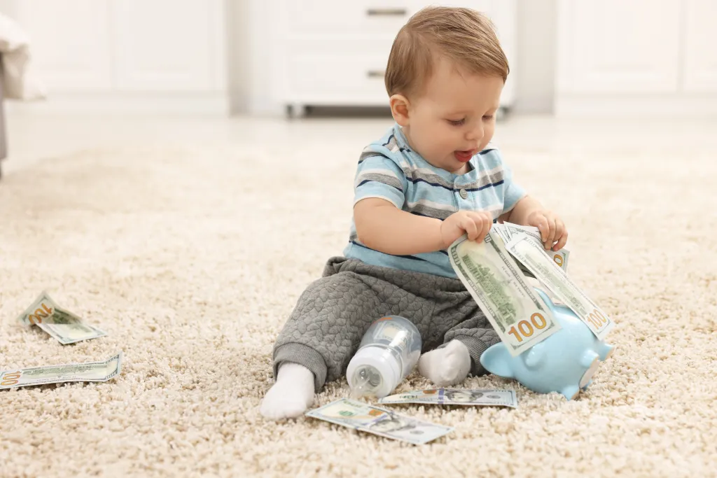 Baby on the floor playing with dollar bills and a piggy bank.