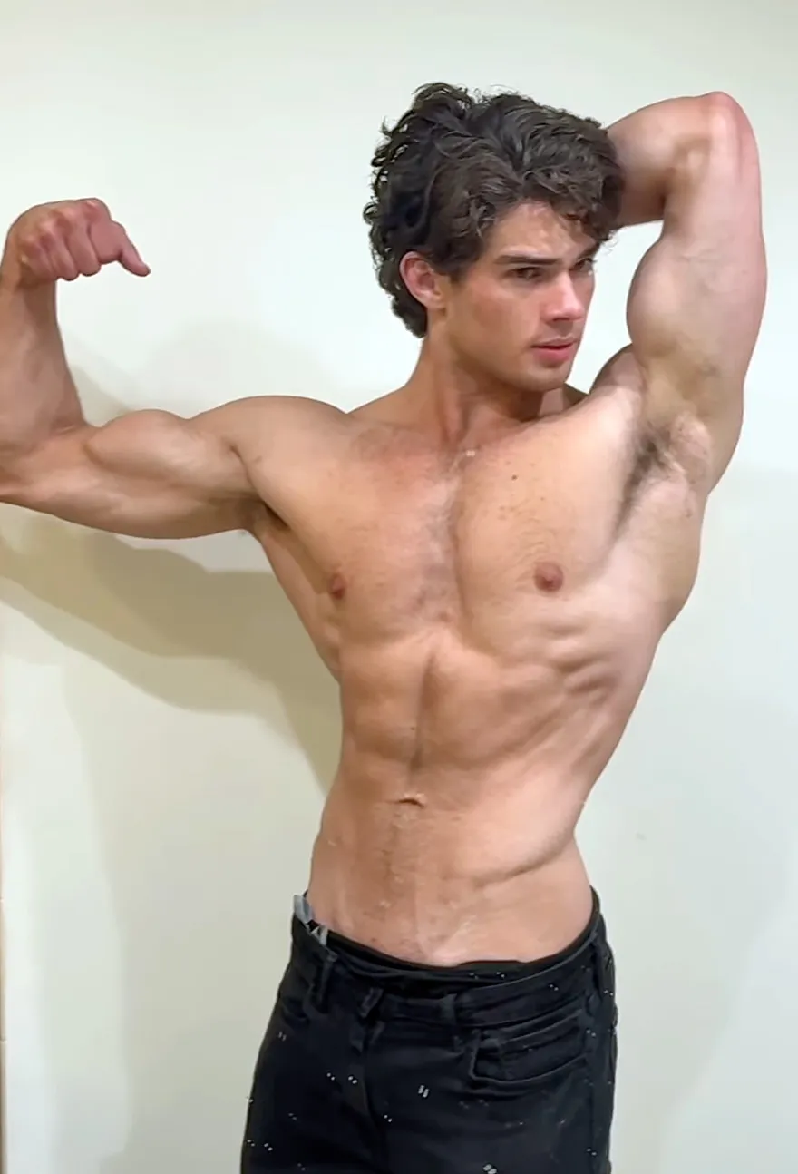 A young muscular man with curly hair and black pants posing and flexing his arm.