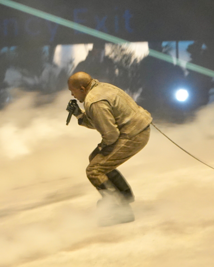 Kanye "Ye" West performing on stage with a microphone in front of a hazy, smoky backdrop.