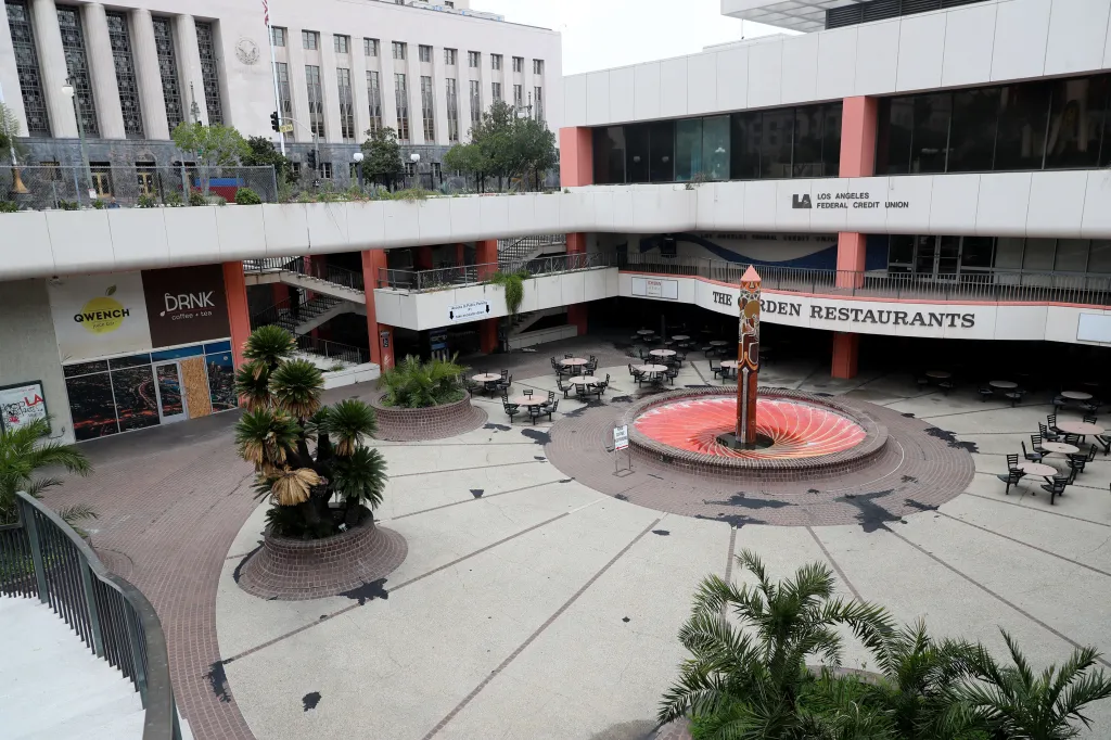 An empty outdoor mall in Los Angeles with vacant storefronts, including Quench Juice Bar and DRNK coffee + tea, and an empty courtyard with tables and chairs.