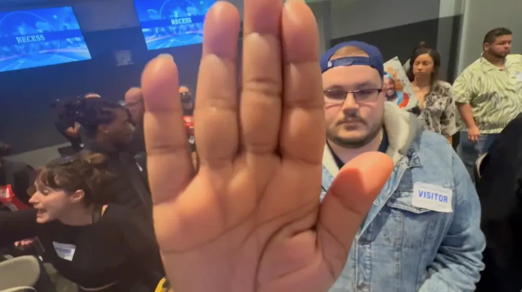 A person holding their hand up to the camera, with a crowd of people and a screen displaying "RECESS" in the background.