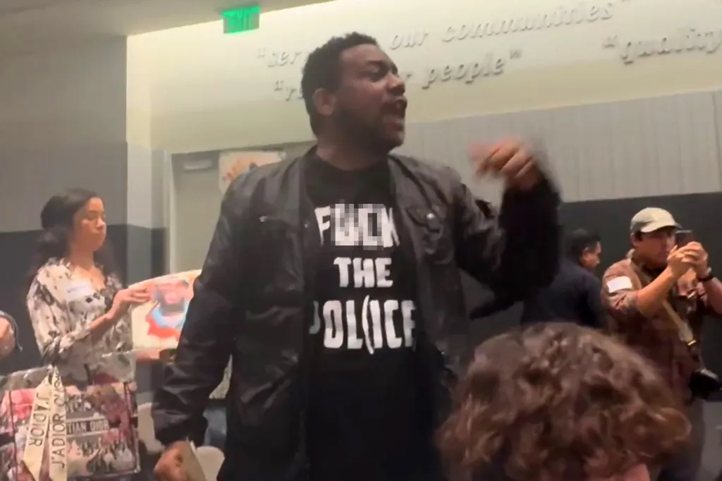 A Black man wearing a black jacket over a black t-shirt that says "FUCK THE POLICE" in white capital letters, with his mouth open as if shouting, at a Los Angeles Police Commission meeting.