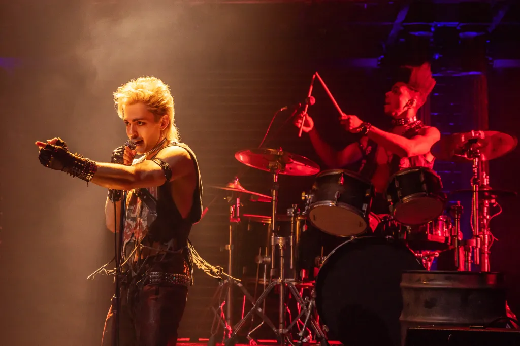 Ali Louis Bourzgui singing while pointing, with Dean Maupin playing drums in the background on the set of "The Lost Boys on Broadway".