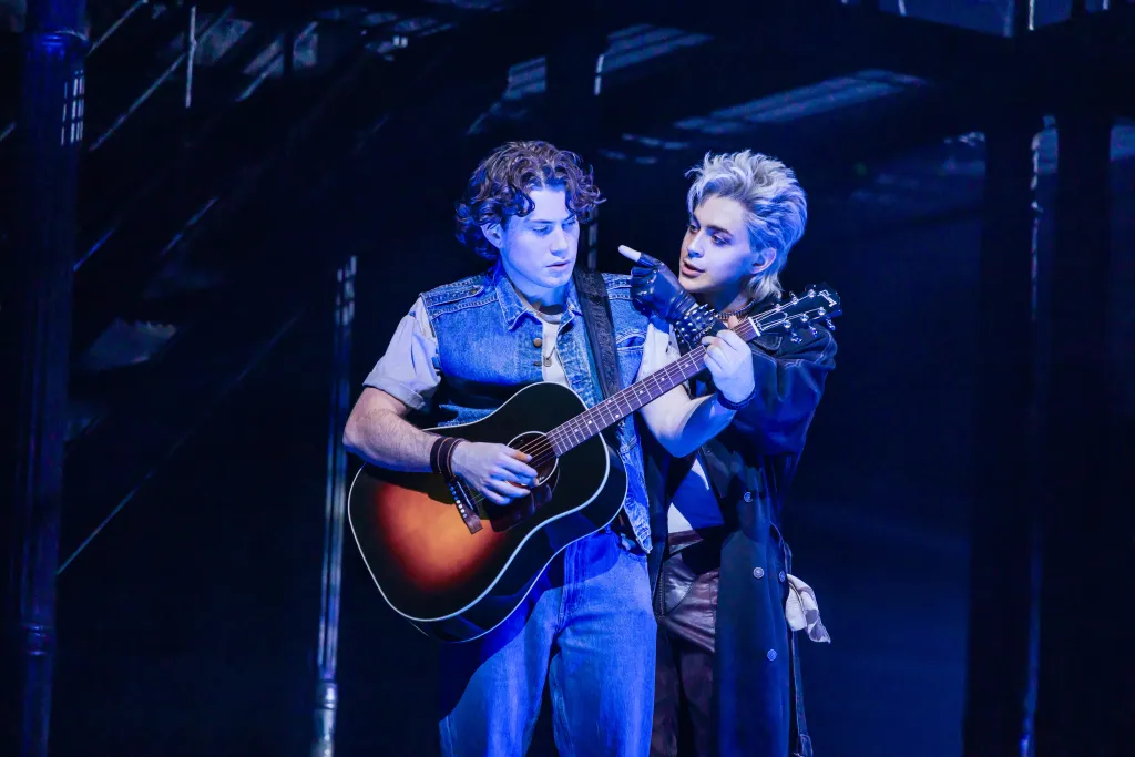 LJ Benet playing an acoustic guitar while Ali Louis Bourzgui points at it, both performing in The Lost Boys on Broadway.