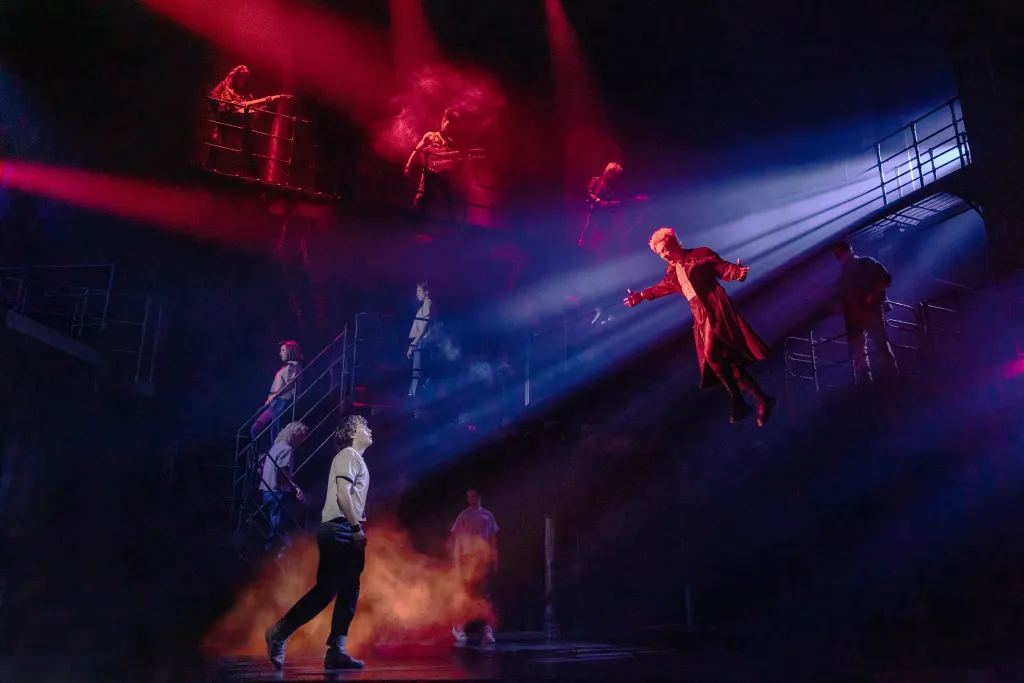 LJ Benet and Ali Louis Bourzgui with the Company perform in "The Lost Boys on Broadway" with several actors on multi-level stage scaffolding and one actor in a long red coat suspended in the air.