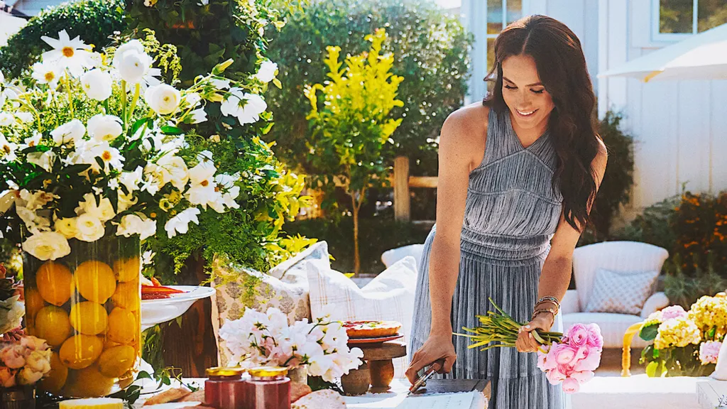 Meghan Markle, in a grey dress, arranging flowers at an outdoor table.
