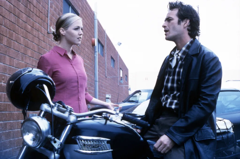 Luke Perry, in a black jacket, blue and white checked shirt and dark pants, and Jennie Garth, in a pink shirt, from the TV show "Beverly Hills, 90210" standing next to a Triumph motorcycle.